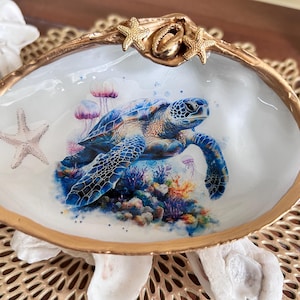 May include: A decorative seashell with a gold rim and starfish accents. The interior features a vibrant watercolor painting of a sea turtle, jellyfish, and coral reef in shades of blue, purple, and orange.