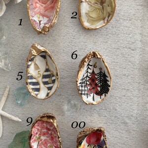 May include: Nine gold oyster shell trinket dishes with different patterns and numbers 1 through 9 and 00. The patterns include a pink rose, green floral, black and white stripes with tassels, red and black plaid trees, pink floral, and red and green floral. The dishes are arranged in a grid pattern on a beige surface.