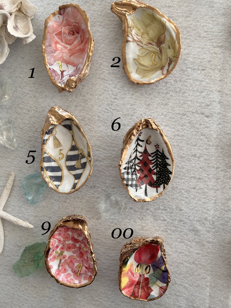 May include: Nine gold oyster shell trinket dishes with different patterns and numbers 1 through 9 and 00. The patterns include a pink rose, green floral, black and white stripes with tassels, red and black plaid trees, pink floral, and red and green floral. The dishes are arranged in a grid pattern on a beige surface.