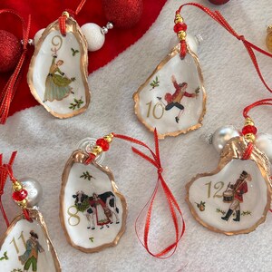 May include: Five oyster shell ornaments featuring gold-painted edges and red ribbon hangers with gold beads. Each shell displays a numbered illustration from the Twelve Days of Christmas, including numbers 8 through 12, with festive holly accents.