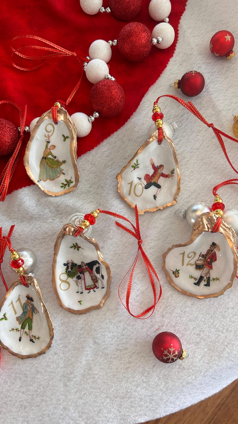 May include: Five oyster shell ornaments featuring gold-painted edges and red ribbon hangers with gold beads. Each shell displays a numbered illustration from the Twelve Days of Christmas, including numbers 8 through 12, with festive holly accents.