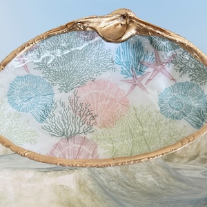 May include: A decorative seashell-shaped dish with a gold-painted rim. The interior features a sea-themed design with coral, seashells, and starfish in pastel colors. The dish is ideal for coastal decor.