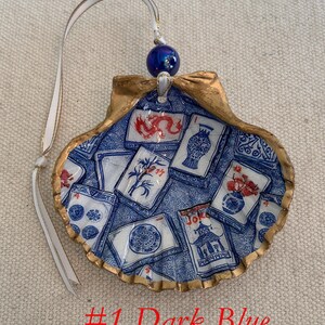 May include: A decorative seashell ornament with a blue and white design, featuring various illustrations. The shell has a gold-colored rim and a white ribbon for hanging. The text "#1 Dark Blue" is visible at the bottom.