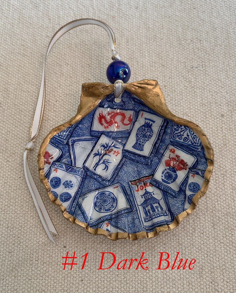 May include: A decorative seashell ornament with a blue and white design, featuring various illustrations. The shell has a gold-colored rim and a white ribbon for hanging. The text "#1 Dark Blue" is visible at the bottom.
