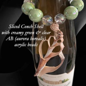 May include: A shell wine bottle charm featuring a sliced conch shell pendant. The charm includes creamy green and clear aurora borealis acrylic beads. The text on the image reads "Sliced Conch Shell with creamy green & clear AB (aurora borealis) acrylic beads" and "Shell Wine Bottle Charm."