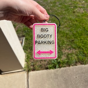 May include: A white sign with pink trim and black text that reads "BIG BOOTY PARKING" with pink arrows pointing left and right. The sign is hanging from a black string.