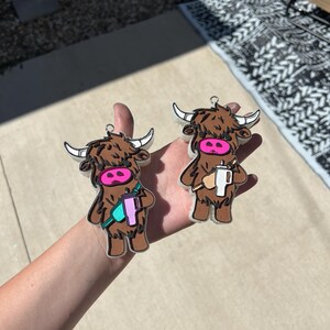 Può includere: Due ornamenti di mucca Highland marroni con nasi rosa e dettagli bianchi. Una mucca tiene una tazza turchese e viola, l'altra tiene una tazza bianca.