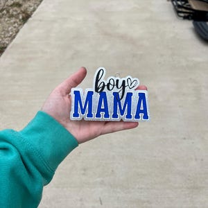 May include: A white sign with blue lettering that says "boy mama" with a small heart symbol after the word "boy".