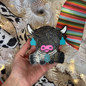 May include: A black and brown Highland cow ornament with turquoise accents and a pink nose. The cow has white horns and is wearing a turquoise necklace and earrings. The ornament is held in front of a white Christmas tree.