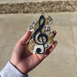 May include: A white, black, and gold glitter Christmas ornament shaped like a treble clef with musical notes surrounding it.