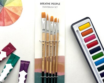 May include: A set of paintbrushes with the text "BREATHE PEOPLE PAINTBRUSH SET". The set includes five paintbrushes with wooden handles and black bristles. Also visible are a watercolor palette and tubes of paint.