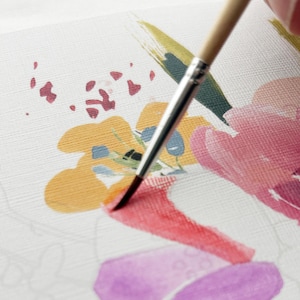 May include: A paintbrush with a light wood handle and silver ferrule applies red paint to a textured white canvas. The canvas features a partially completed floral illustration with yellow, pink, purple, and dark blue painted shapes and outlines.
