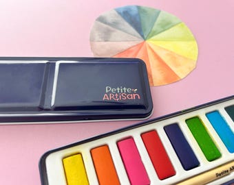 May include: A dark blue metal watercolor paint tin with the text Petite Artisan printed in pink and green. The set includes a white plastic tray with rectangular paint pans in yellow, orange, pink, red, purple, green, and blue, plus a color wheel.