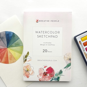 May include: A watercolor sketchpad with the text "WATERCOLOR SKETCHPAD" and dimensions of 6 x 8 inches. A watercolor palette with a paintbrush and a watercolor color wheel are also present. The sketchpad has floral illustrations.