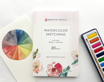 May include: A watercolor sketchpad with the text "WATERCOLOR SKETCHPAD" and dimensions of 6 x 8 inches. A watercolor palette with a paintbrush and a watercolor color wheel are also present. The sketchpad has floral illustrations.