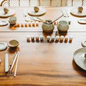 May include: A wooden table with pottery supplies, including clay, paint, brushes, and tools. There are two small bowls, two plates, and two small containers of paint.
