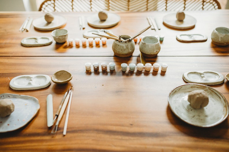 May include: A wooden table with pottery supplies, including clay, paint, brushes, and tools. There are two small bowls, two plates, and two small containers of paint.