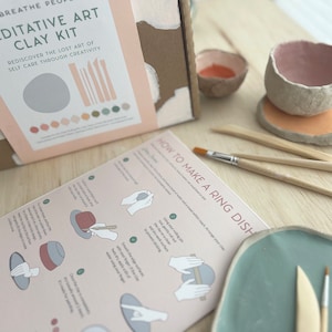 May include: A craft kit for making clay art. The box has a pink and brown design with the text "Meditative Art Clay Kit" and "Rediscover the lost art of self care through creativity". The kit includes tools, clay, and instructions for making a ring dish.