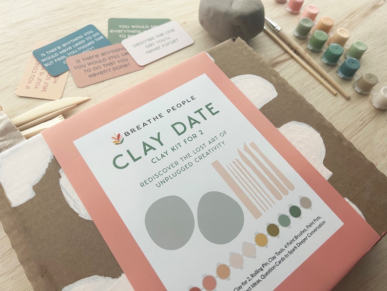 May include: A pink and brown box with the text "BREATHE PEOPLE CLAY DATE" and "CLAY KIT FOR 2". The box contains clay, tools, paint, and conversation cards for a creative date night.