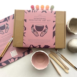 May include: A craft kit for meditative art, featuring a cardboard box with a pink label that reads "Honoring Meditative Art Kit." The kit includes clay, paint, brushes, and tools. A pink bowl and two clay bowls are also visible.