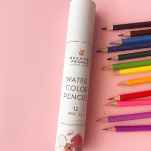 May include: A white cylindrical cardboard tube labeled BREATHE PEOPLE WATERCOLOR PENCILS 12 PENCILS with a floral illustration at the base. Twelve sharpened watercolor pencils in various colors are arranged in a fan shape next to the tube.