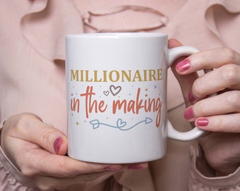 May include: White ceramic mug with the text "MILLIONAIRE in the making" in gold and red lettering. The mug features hearts, stars, and an arrow design. The mug is held by a person with pink nail polish.