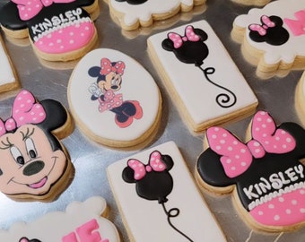 May include: A collection of decorated sugar cookies featuring a cartoon mouse character with a pink bow. Some cookies have the word "ONE" in pink icing, while others have the name "Kinsley" in pink icing.