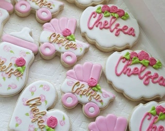 May include: A set of nine decorated sugar cookies with pink and white frosting. The cookies feature the name "Chelsea" and images of baby carriages and a baby bottle.