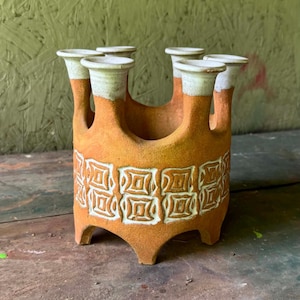 May include: A terracotta ceramic flower frog with five individual tubular openings. The vessel features a matte orange finish with a band of white embossed square geometric patterns around the center and white rims on each of the five top openings.