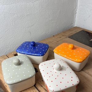 Könnte beinhalten: Vier Keramik-Butterdosen mit Deckeln. Die Deckel sind blau, orange, weiß und grün, jeweils mit einem Tupfenmuster. Die Dosen sind rechteckig mit weißem Boden und einem Knopf oben.