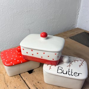 Könnte beinhalten: Drei Keramik-Butterdosen mit Deckel. Eine ist weiß mit roten Punkten, eine andere ist weiß mit roten Herzen, und die dritte ist weiß mit dem Wort "Butter" und einem Astmuster. Jede hat einen Knopf oben.