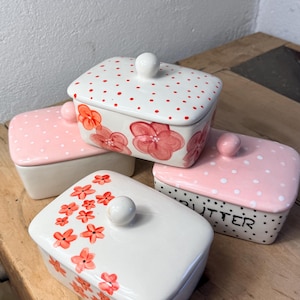 Könnte beinhalten: Eine Sammlung von Keramik-Butterdosen mit Deckel. Die Dosen sind weiß, rosa und schwarz und mit roten Punkten und floralen Mustern verziert. Eine Dose trägt das Wort "BUTTER" in Schwarz.