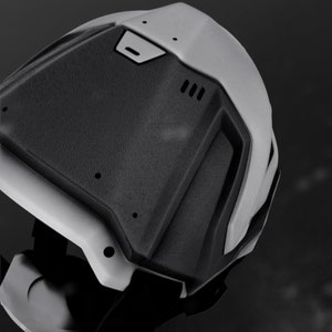 May include: A black and gray helmet with a textured surface and a raised design on the back. The helmet has a small, rectangular vent on the top and a larger, rectangular vent on the back.