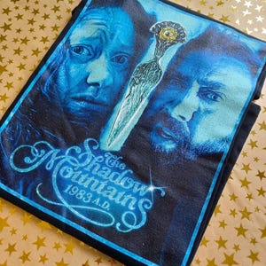 May include: Black t-shirt with a blue graphic of two faces and a dagger. The text "The Shadow Mountains 1983 A.D." is in a light blue, glittery font. The faces are in shades of blue.