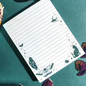May include: A white lined notepad with a green butterfly, a bird, a cat, and feathers on the edges. The notepad is on a teal background with various stones and feathers.