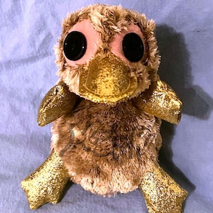 May include: A plush platypus toy featuring shaggy brown fur, large black button eyes, and a metallic gold fabric beak. The creature has matching glittery gold fabric feet and flippers, with a soft, rounded body and a textured, fuzzy appearance.