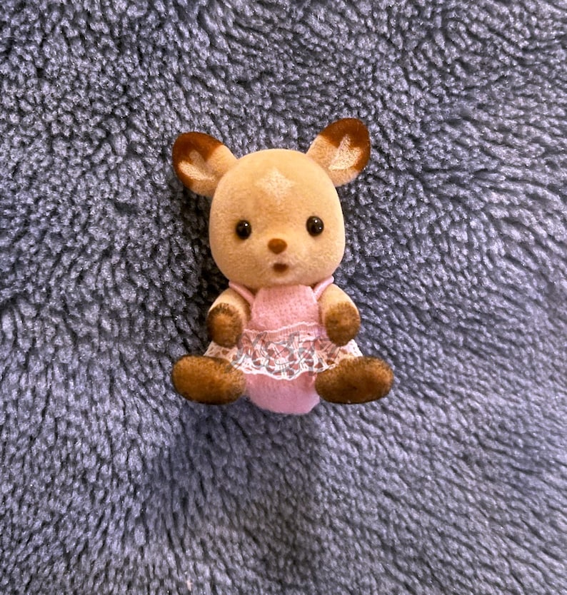 以下が含まれることがあります： 耳、足、手に茶色のアクセントが付いた、小さな薄茶色の鹿の置物。白いレースのトリムが付いたピンク色のドレスを着ています。置物は黒い目をし、驚いた表情をしています。