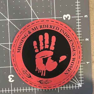 May include: A circular red and black patch featuring a red handprint graphic in the center and the text "MISSING & MURDERED INDIGENOUS WOMEN" printed along the border. Two small feather icons are at the bottom. It measures 3 inches in diameter.