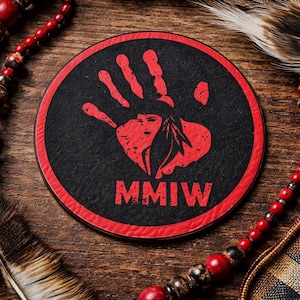 May include: A round black coaster with a red border. The coaster features a red handprint design with a Native American headdress and the letters MMIW. The coaster is on a wooden surface with decorative beads and feathers.