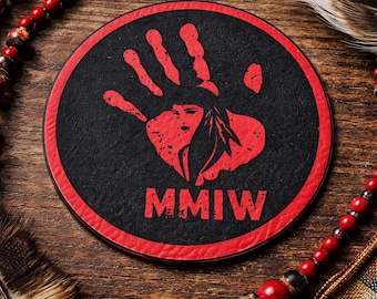 May include: A round black coaster with a red border. The coaster features a red handprint design with a Native American headdress and the letters MMIW. The coaster is on a wooden surface with decorative beads and feathers.