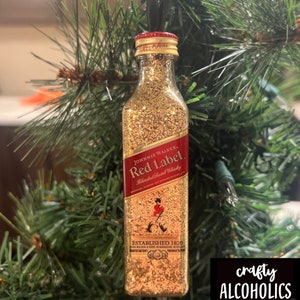 Puede incluir: Adorno de botella de purpurina dorada con una etiqueta roja que dice "Johnnie Walker Red Label Blended Scotch Whisky Established 1820".