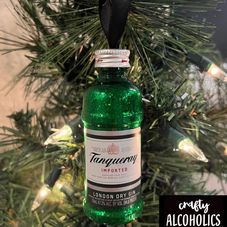 May include: A green glittered Tanqueray gin bottle Christmas ornament. The bottle has a white label with black text and a silver cap. The ornament hangs from a black ribbon and is displayed on a Christmas tree.