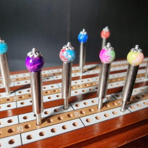 May include: A close-up of a wooden cribbage board with nine colorful, decorative pegs. The pegs are made of glass and have a silver crown on top. The pegs are inserted into the holes on the cribbage board.