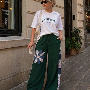 Puede incluir: Una camiseta blanca con la inscripción "ANINE BING SPORT" en verde, combinada con pantalones anchos de color verde oscuro. Los pantalones presentan una aplicación floral y paneles laterales con estampado floral. El conjunto se completa con zapatillas y gafas de sol.