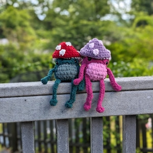 May include: Two crocheted mushroom-shaped plushies sitting on a grey wooden fence. The mushroom on the left is teal with a red and white cap. The mushroom on the right is pink with a purple and white cap.