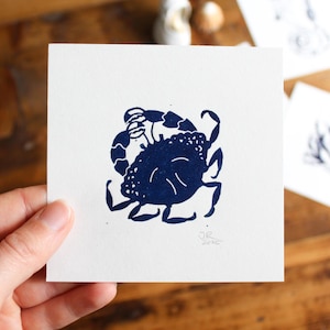 Pode incluir: Uma gravura em linóleo azul marinho de um caranguejo em um cartão quadrado branco. O caranguejo é retratado em um design estilizado e minimalista. As iniciais do artista e o ano de 2020 são visíveis no canto inferior direito. O cartão é segurado por uma mão.