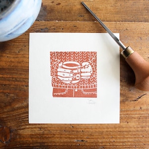 Op de afbeelding: Een vierkante, rood-oranje linoleumsnede op wit papier toont een persoon die een mok vasthoudt. De persoon draagt een trui en een spijkerbroek. Een snijgereedschap en een keramische kom staan op de achtergrond. De prent staat op een houten oppervlak.