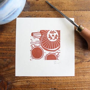 Op de afbeelding: Een vierkante print met een rood-oranje linoleumsnede ontwerp van een theepot, kopjes en een decoratief element. Een snijgereedschap met een houten handvat en metalen mes ligt op het houten oppervlak. Een blauw-witte keramische kom bevindt zich in de linkerbovenhoek.