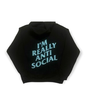 Puede incluir: Sudadera con capucha negra con el texto "I'M REALLY ANTI SOCIAL" en azul claro en la espalda. La sudadera tiene capucha y mangas largas. El fondo es blanco.