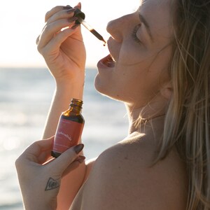 May include: A woman holding a small amber glass bottle and dropper, with the label "Aphrodisia". The woman is tilting her head back and opening her mouth, as a liquid drips from the dropper. The background is a blurred view of the ocean.